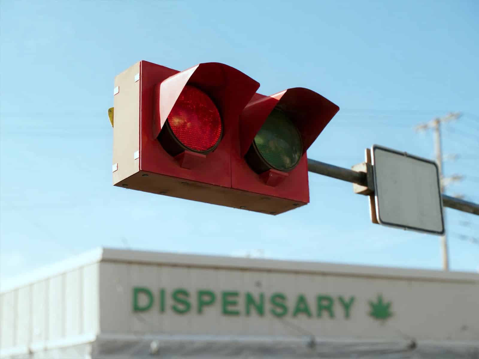 Rot leuchtende Ampel vor hellem, blauem Tageshimmel, im Hintergrund ein Dispensary-Schild mit Cannabisblatt-Logo – Sinnbild für die Ampel-Matrix im Werbeverbot